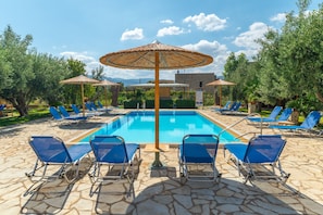 Un área de piscina con sillas reclinables azules y sombrillas de paja, rodeada de pavimento de piedra y follaje.