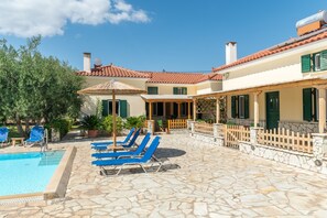 Un espace piscine avec des chaises longues, une terrasse en pierre et un bâtiment doté d’un toit en tuiles.