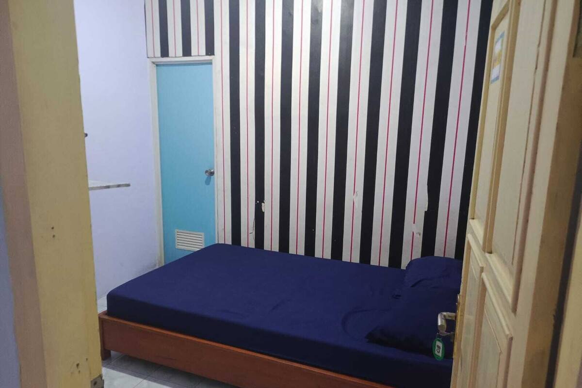 A small, single-bed room with a striped wall, a blue door, and a wooden bed frame.