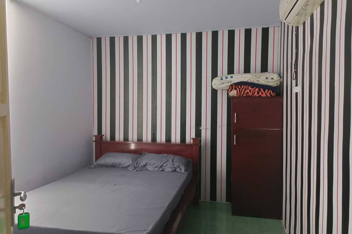 A single-bed room with a striped wall, a ceiling fan, an air conditioning unit, and a green carpet.