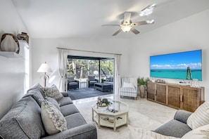 Een moderne woonkamer met een grote televisie, comfortabele zitplaatsen, een plafondventilator en uitzicht op de oceaan.
