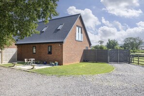A single-story brick house with a sloped roof, a small patio with outdoor furniture, and a gravel driveway. - Beautiful Self Contained Annex in Toddington (Cheltenham)