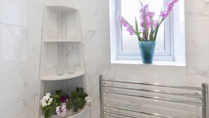 A bathroom with white marble walls, a window with a view, a shelf with flowers, and a towel rack. - Beautiful Self Contained Annex in Toddington (Cheltenham)