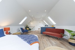 A modern loft bedroom with a large bed, a red sofa, a blue armchair, a small kitchen area, and skylights.