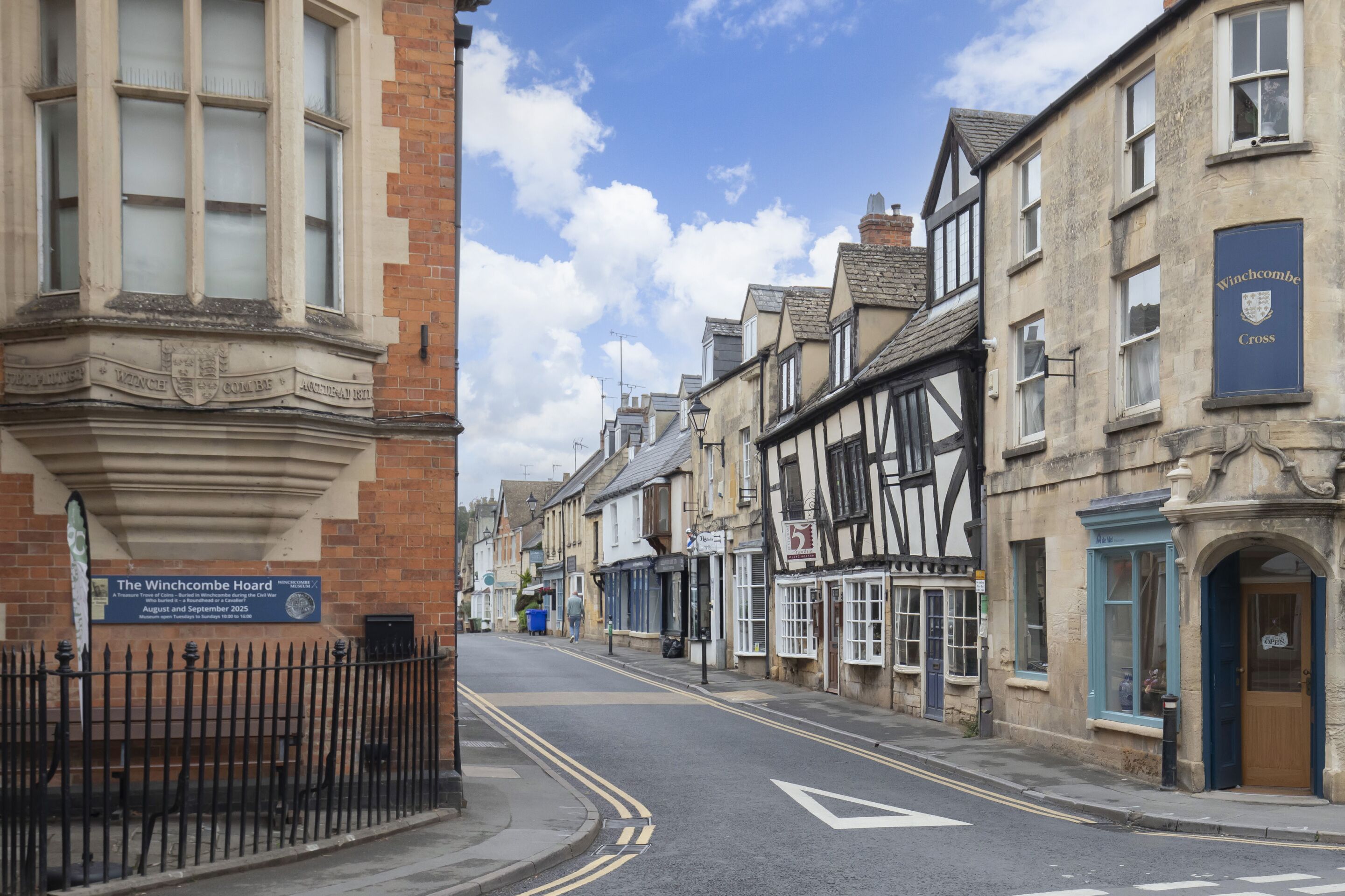 Rua com edifícios tradicionais, céu claro e uma placa para The Winchcombe Hoard.