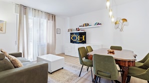 A modern living room with a dining table, green chairs, a white sofa, a TV on the wall, and a bookshelf.