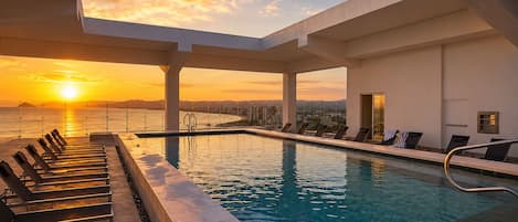 Zona de piscina con tumbonas y vistas a la puesta de sol sobre el océano.