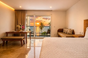 A hotel room with a bed, a desk with a chair, a balcony with a view of the sunset over the ocean, and a sofa.