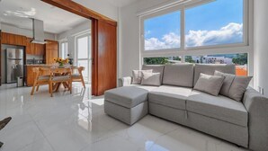 A modern living room with a grey sectional sofa, wooden dining table, and a kitchen area with wooden cabinets. - Amazing 1BR - Rooftop Pool in Versalles (Puerto Vallarta)