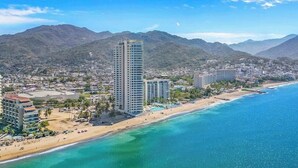 Un paisaje costero con un hotel moderno y alto destacándose, una playa y montañas al fondo.