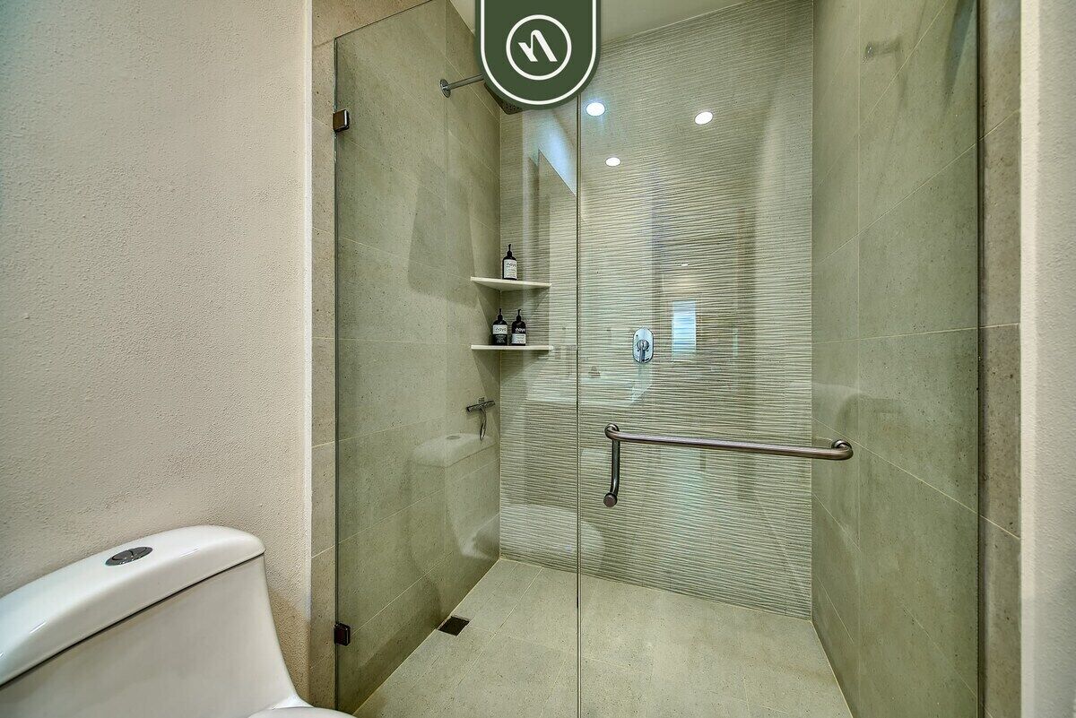 A glass-enclosed shower stall with a handrail, a toilet, and a shelf with toiletries.