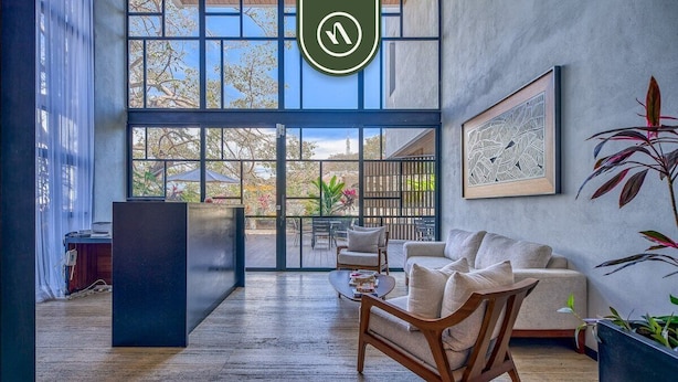 A modern interior with a large window, a framed artwork, and a wooden chair. - New 2BR Condo in Zona Romantica - Rooftop Pool (Puerto Vallarta)