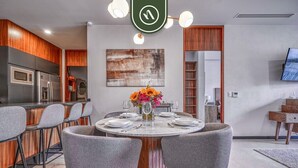 A modern kitchen with a built-in microwave and refrigerator, a dining area with a round table and grey chairs, and a TV mounted on the wall.