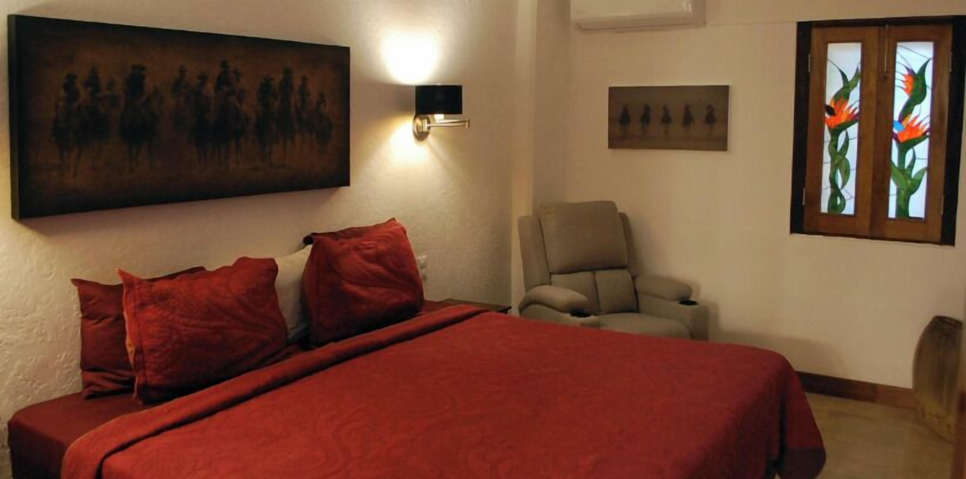 A bedroom with a bed, pillows, a chair, a wall-mounted light, and artwork.