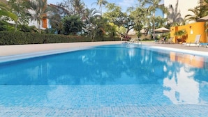 Une piscine d’un bleu éclatant, dont le fond est recouvert de carreaux, entourée de palmiers et bordée d’un bâtiment à la façade jaune.