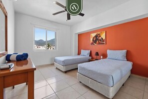 Quarto de hotel com duas camas, uma escrivaninha, ventoinha no teto, janela com vista para as montanhas e uma parede de destaque laranja.