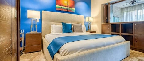 Una camera d'albergo con un letto grande, un comodino, una scrivania, una sedia, un quadro appeso al muro e una ventola a soffitto.