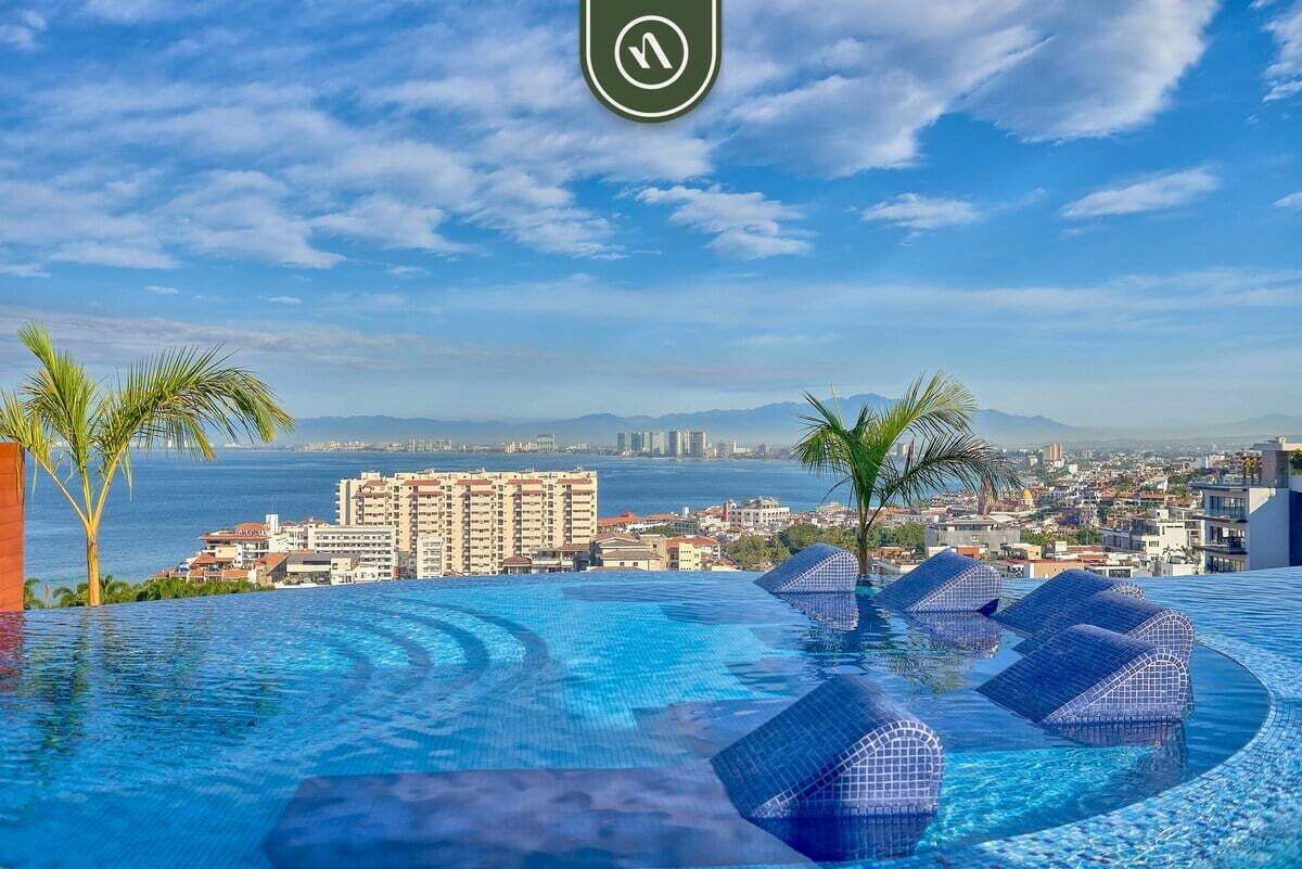 A pool with clear blue water, palm trees, and a view of a cityscape with buildings and mountains in the distance.