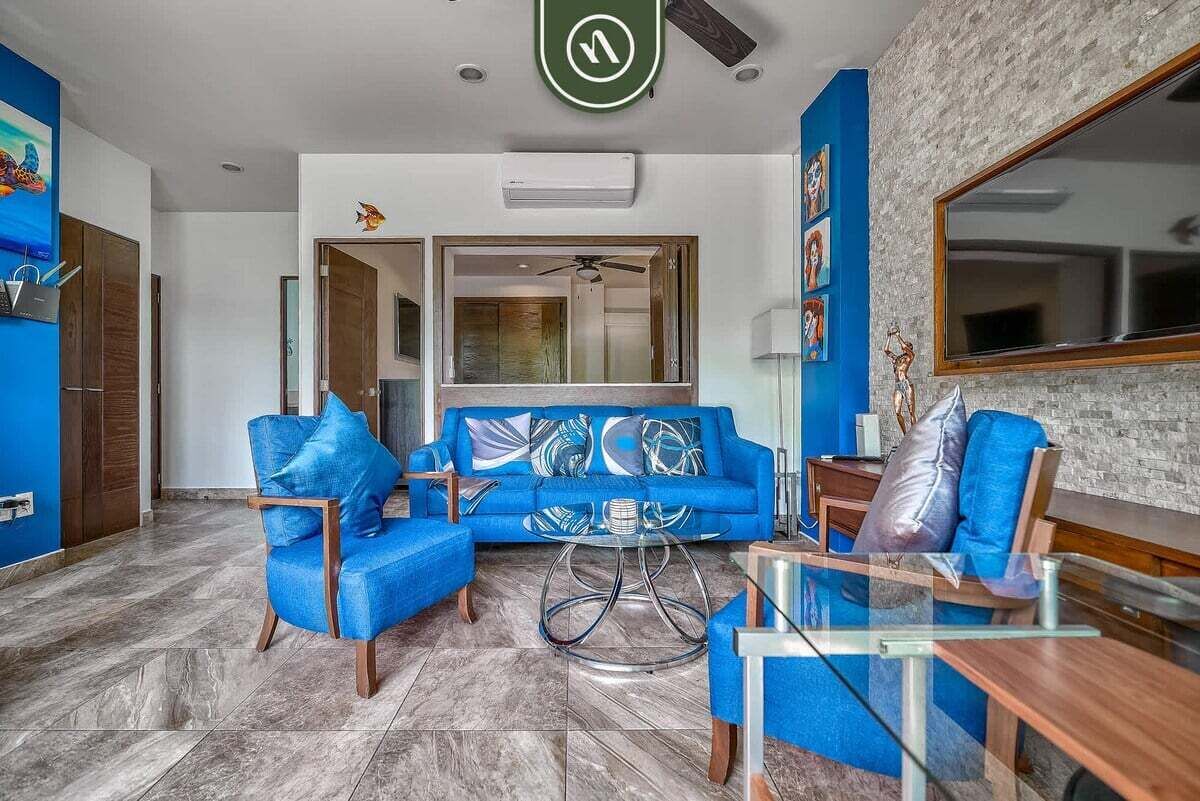 A modern living room with a blue sofa, matching armchairs, a glass coffee table, and a large mirror.