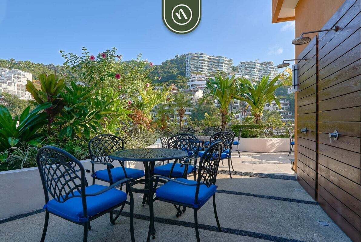 An outdoor seating area with metal chairs and tables, surrounded by greenery and overlooking a cityscape.