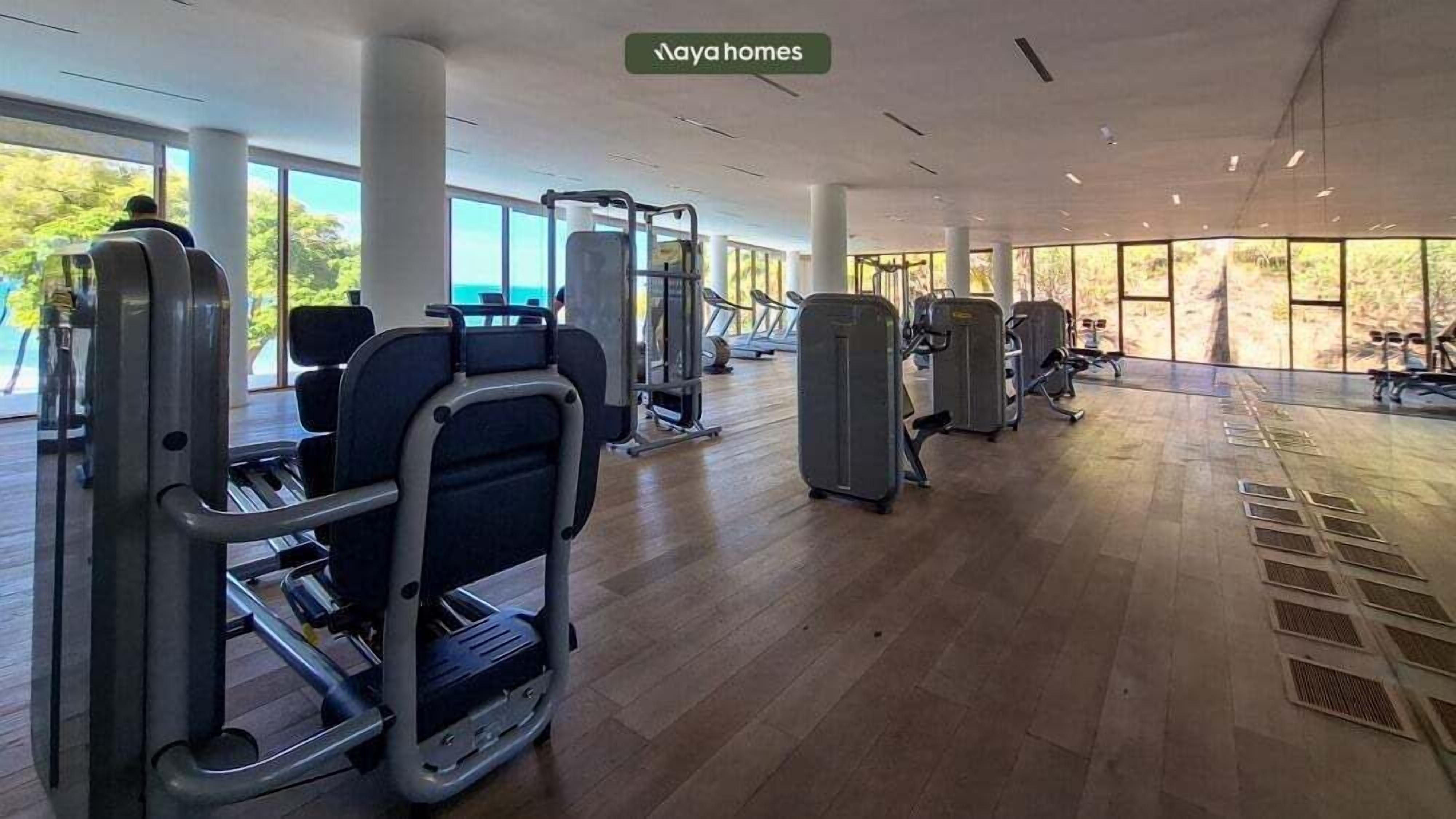 Une salle de sport spacieuse, équipée de divers appareils d’exercice, dotée de grandes fenêtres et d’un plancher en bois.