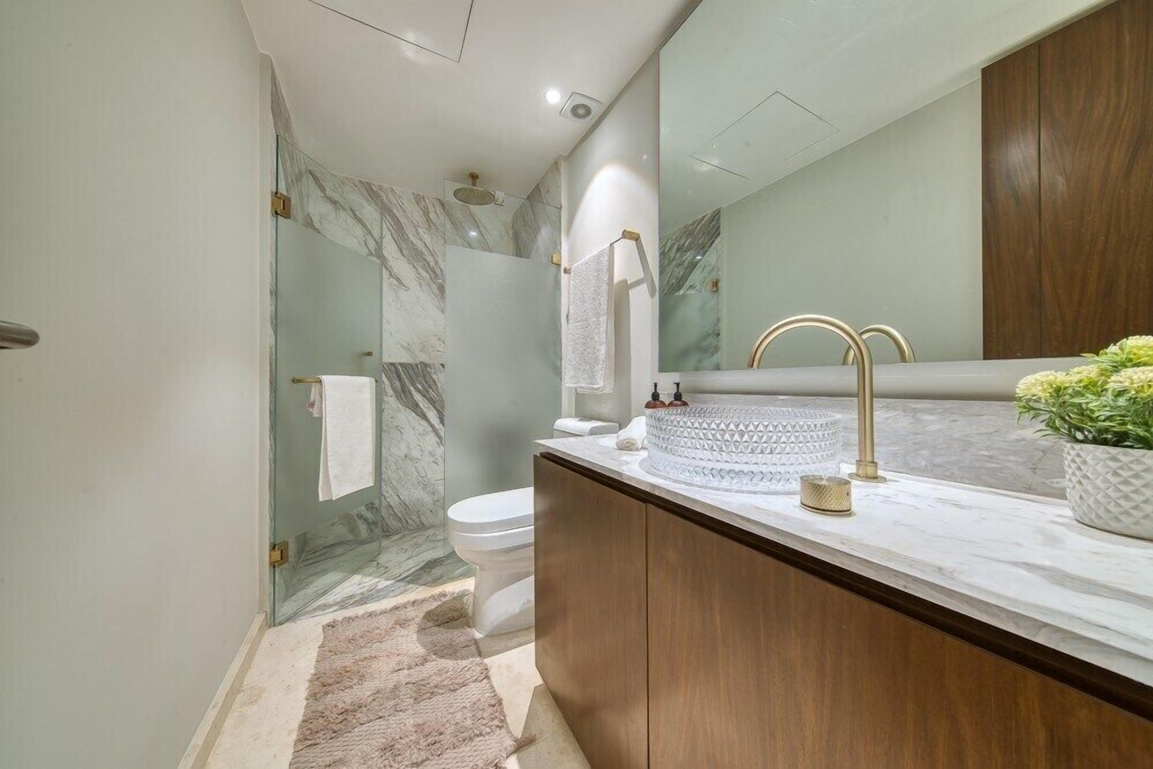 Un baño con ducha de mármol, inodoro, lavamanos con grifo dorado y un espejo grande.