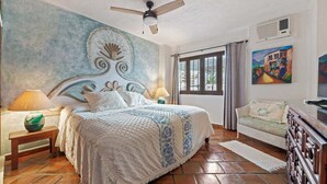 Een slaapkamer met een bed, een plafondventilator, een nachtkastje, een lamp, een schilderij en een raam.