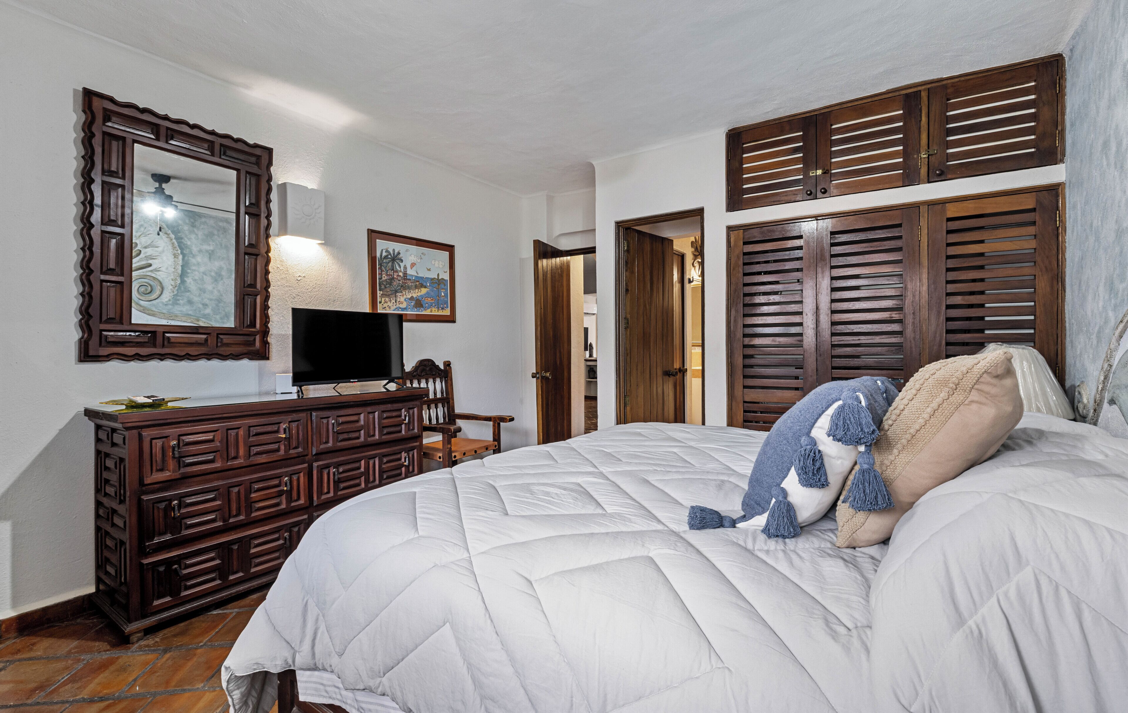 A bedroom with a large bed, wooden wardrobe, a dresser, a television, and a door leading to another room.