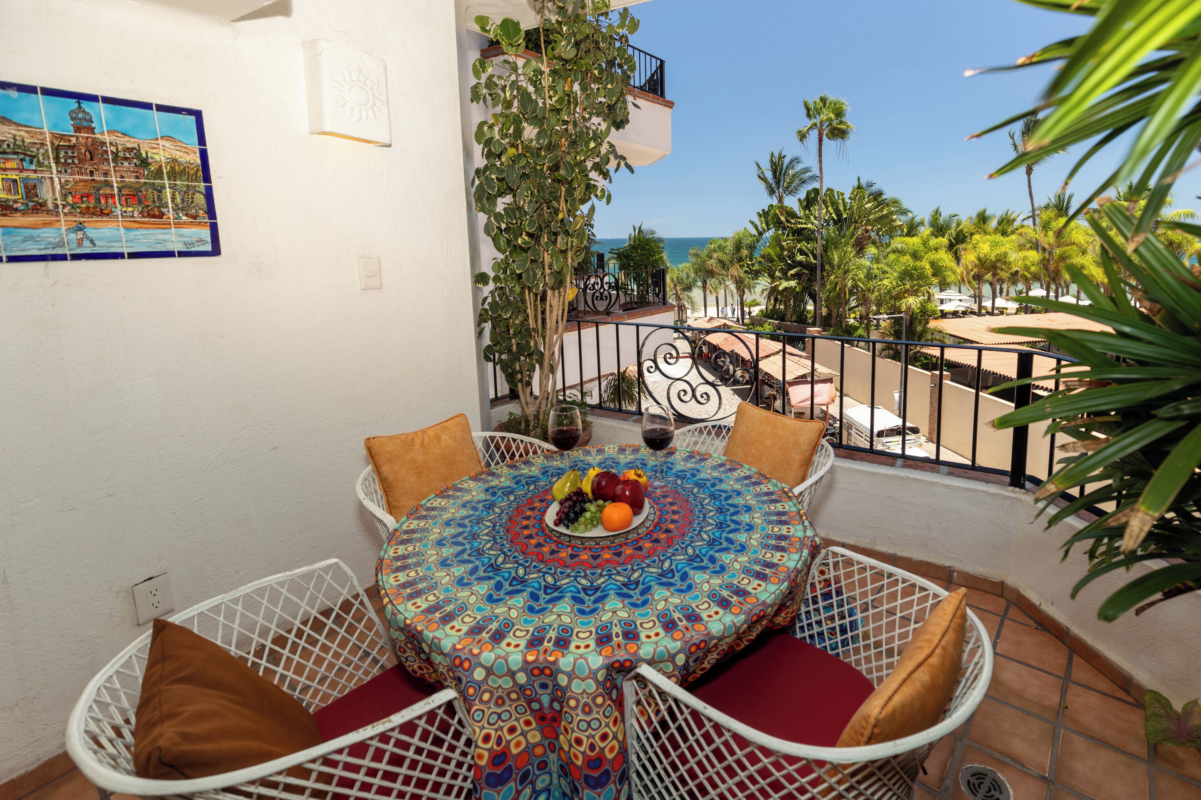 Een ronde tafel met een kleurrijke tafelkleed, omringd door witte stoelen met rode kussens, op een buitenbalkon met uitzicht op zee en palmbomen.