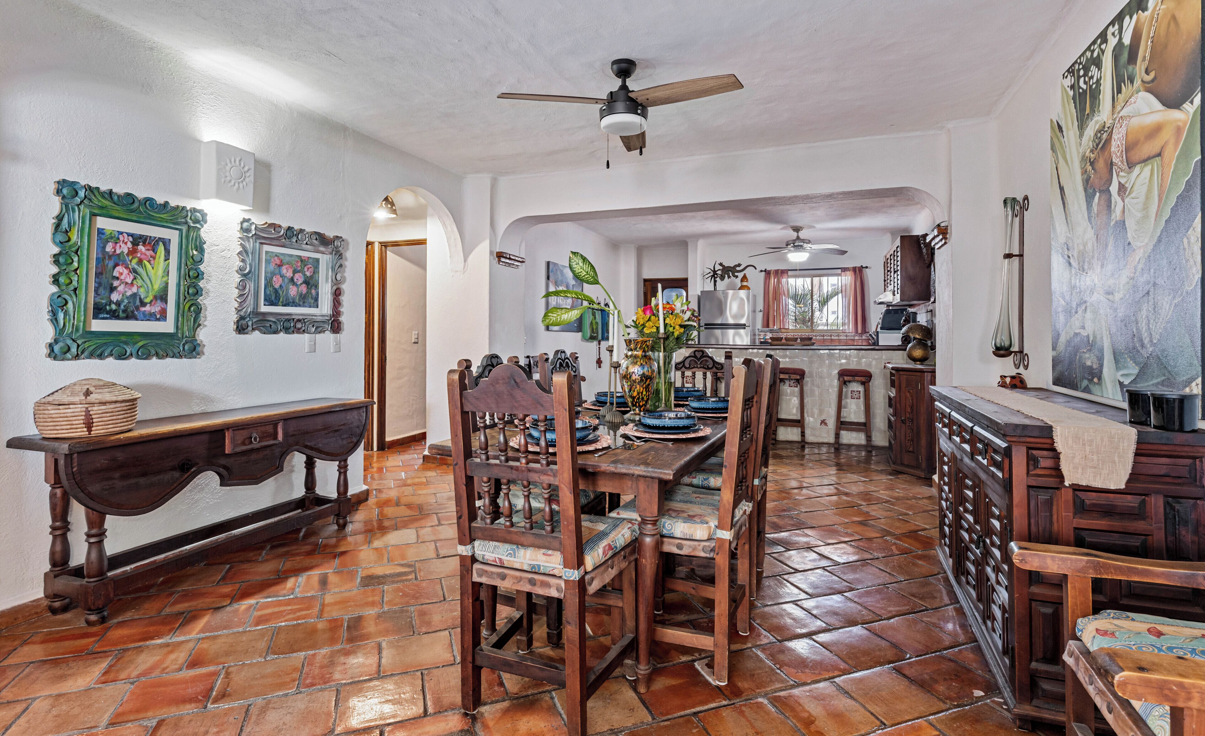 Un coin repas avec une table et des chaises en bois, un ventilateur de plafond et une cuisine en arrière-plan.