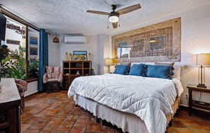 A bedroom with a large bed, a ceiling fan, a window with curtains, a wooden nightstand, and a wall-mounted air conditioner.