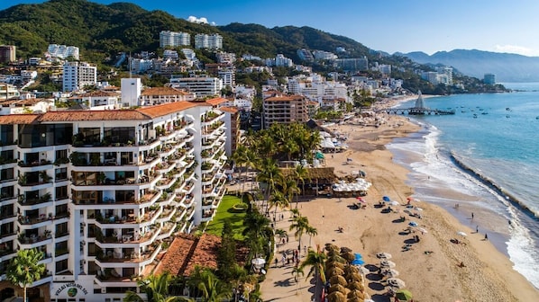 A coastal cityscape with a beach, a prominent hotel with multiple balconies, and a mountainous backdrop.