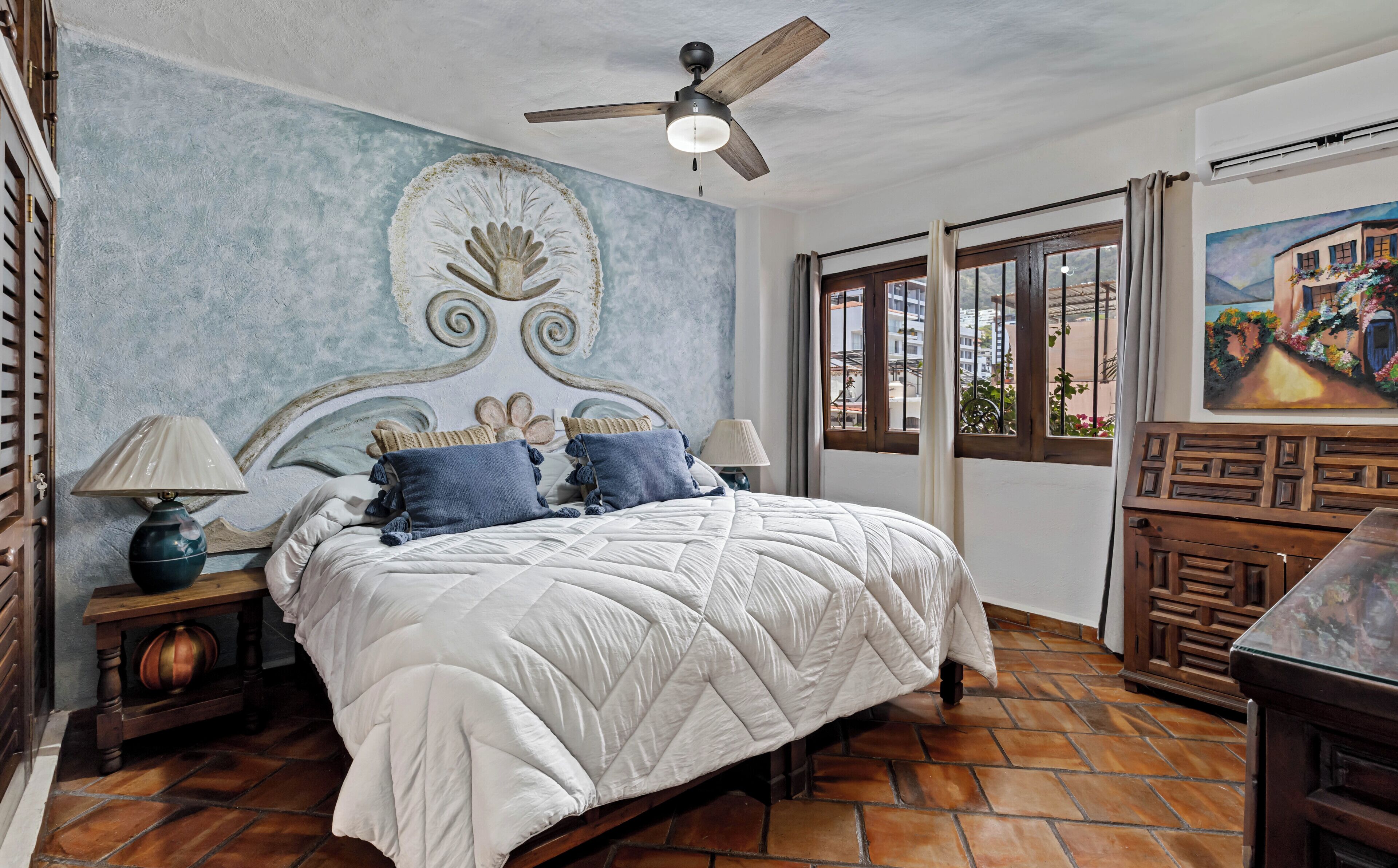 A bedroom with a bed, a ceiling fan, a nightstand, a lamp, a window with curtains, and a painting on the wall.