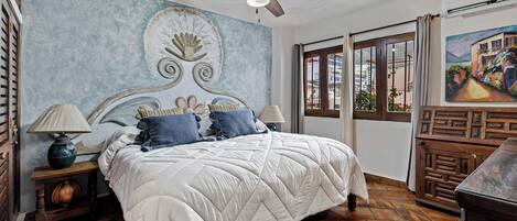 A bedroom with a bed, a ceiling fan, a nightstand, a lamp, a window with curtains, and a painting on the wall.