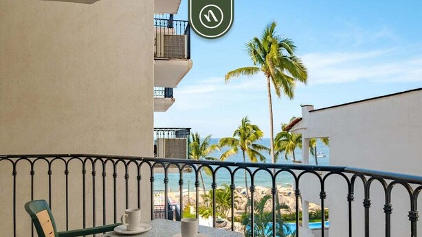 A balcony with a table and chairs, overlooking a beach and a pool. - Amazing 1 BR Condo - Partial Oceanview - Pool (Puerto Vallarta)