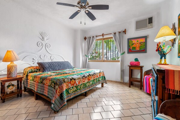 A room with a bed, a ceiling fan, a window with curtains, a wall with a painting, and a lamp.