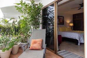 Ein Balkon mit Topfpflanzen, ein Sessel mit Kissen und ein Bett mit einem gelben Kopfkissen im angrenzenden Zimmer.