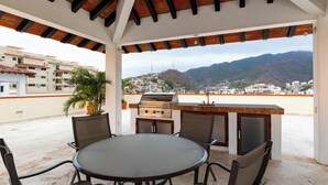 An outdoor patio with a round table and chairs, a grill, and a view of a cityscape and mountains. - Ocean View 2Br Condo-balcony-beach-pool - 902 (Puerto Vallarta)