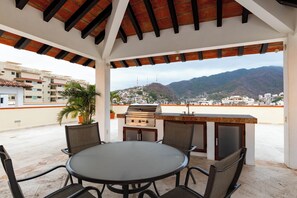Une terrasse extérieure avec une table ronde et des chaises, un barbecue et une vue sur le paysage urbain et les montagnes.