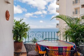 Een ronde tafel met een kleurrijk tafelkleed, omringd door metalen stoelen, op een balkon met potplanten en uitzicht op de zee.