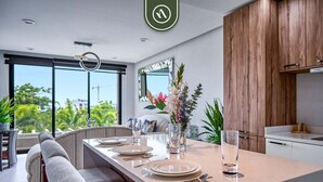 A modern kitchen with wooden cabinets, a dining area with a long table set for four, and large windows offering a view of greenery outside. - 2BR Condo in Bucerias - Pickleball Court - Gym (Bucerías)