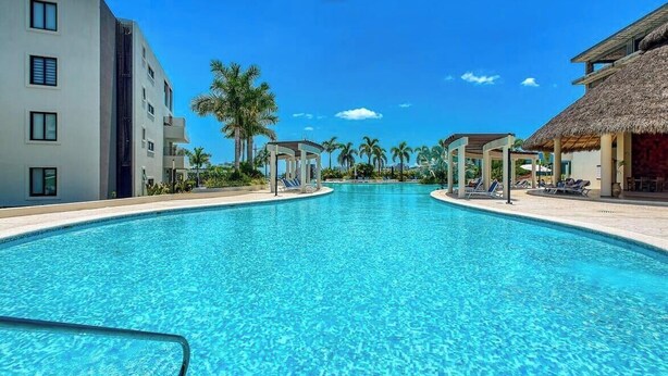 A clear blue pool with lounge chairs and a thatched-roof structure, surrounded by palm trees and modern buildings. - 2BR Condo in Bucerias - Pickleball Court - Gym (Bucerías)