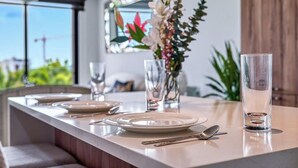 A modern dining area with a long table set for four, clear glasses, a vase with flowers, and a view of greenery outside. - 2BR Condo in Bucerias - Pickleball Court - Gym (Bucerías)