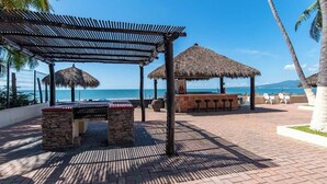 A beachside patio with a thatched-roof structure, a stone bench, and a bar area. - Beachfront 2BR Condo - Pool - Blue Flag Beach (Nuevo Nayarit)