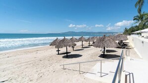 A beach with thatched umbrellas, a railing, and a view of the ocean and distant mountains. - Beachfront 2BR Condo - Pool - Blue Flag Beach (Nuevo Nayarit)