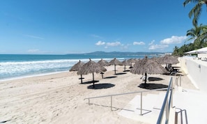 A beach with thatched umbrellas, a railing, and a view of the ocean and distant mountains.