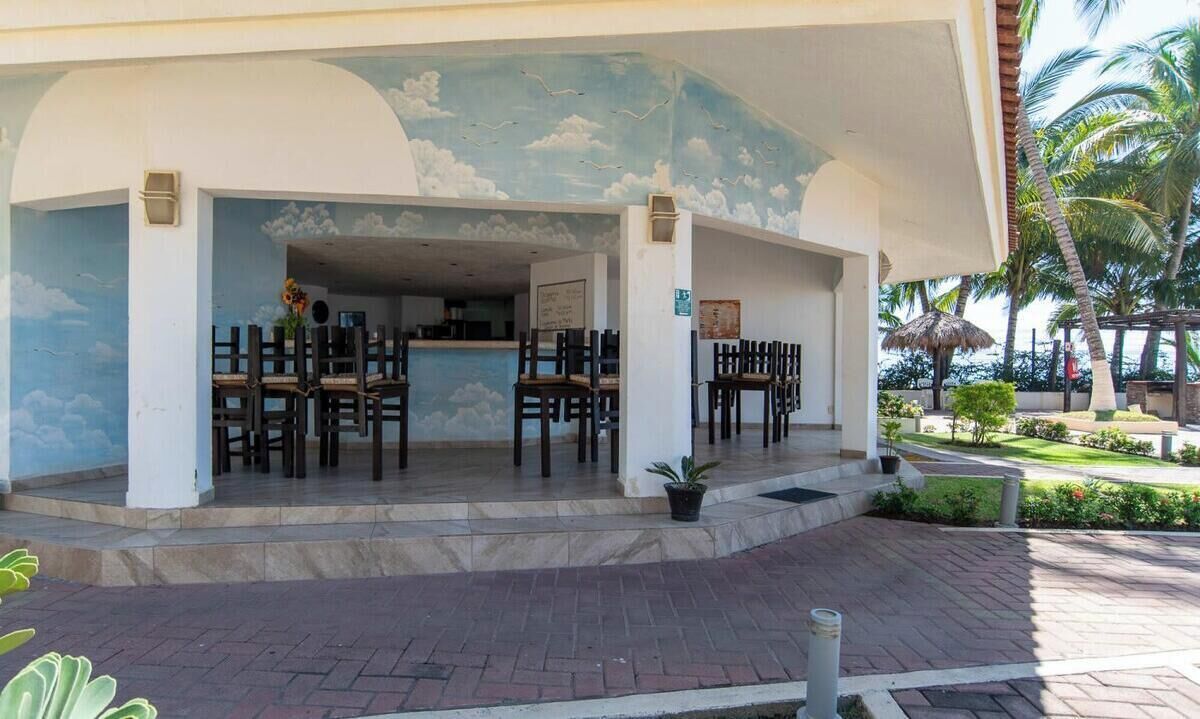 A covered outdoor seating area with chairs and a counter, surrounded by a landscaped garden with palm trees.