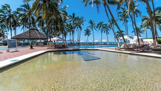 A clear pool with palm trees, lounge chairs, and a thatched-roof structure. - Beachfront 2BR Condo - Pool - Blue Flag Beach (Nuevo Nayarit)