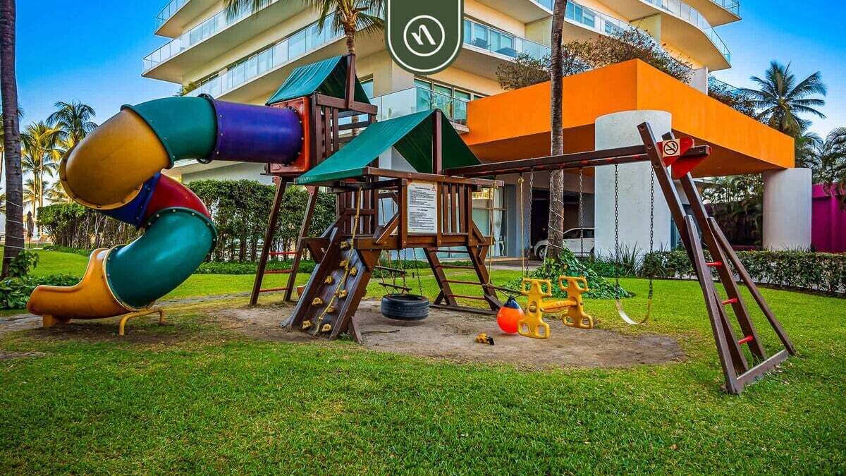 Ein Spielplatz mit Rutsche und Schaukeln vor einem Gebäude mit Palmen und blauem Himmel.