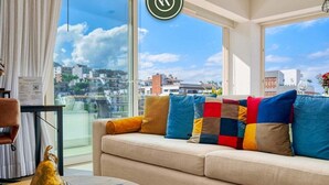 A modern living room with a beige sofa, colorful cushions, a glass coffee table, and a view of a cityscape through large windows.
